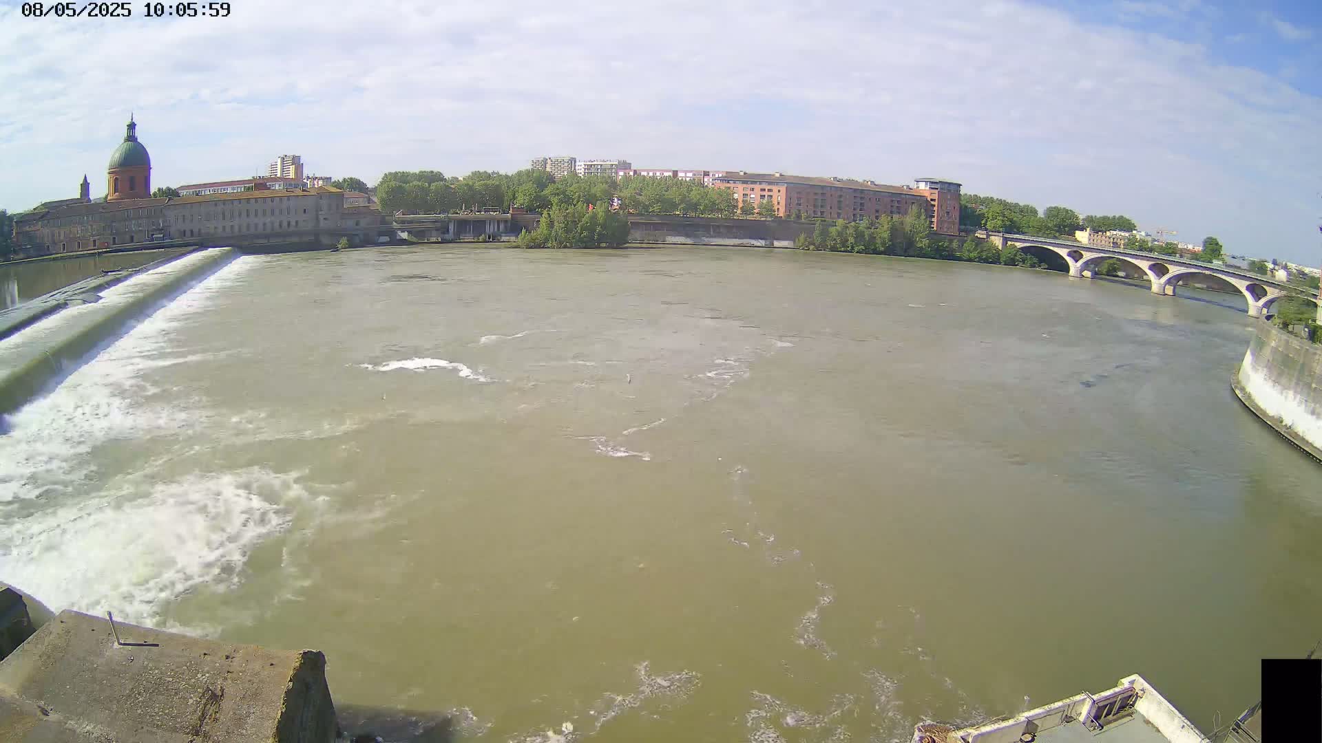 A wide, muddy river flows under a bridge, past a dam, and toward buildings under a partly cloudy sky.