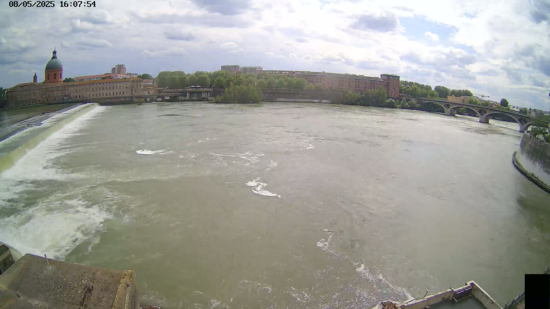A wide shot of a river with a dam and buildings in the background under a partly cloudy sky.