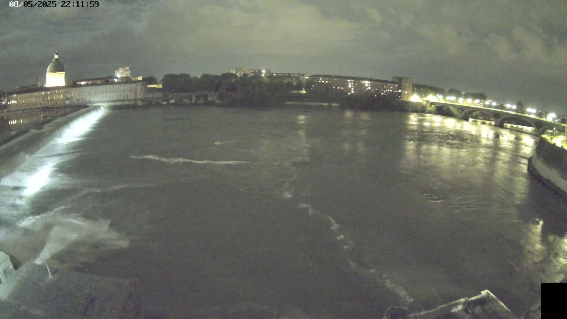 A nighttime, overcast view shows a river flowing past a city with illuminated buildings and a bridge, with a dam visible in the foreground.