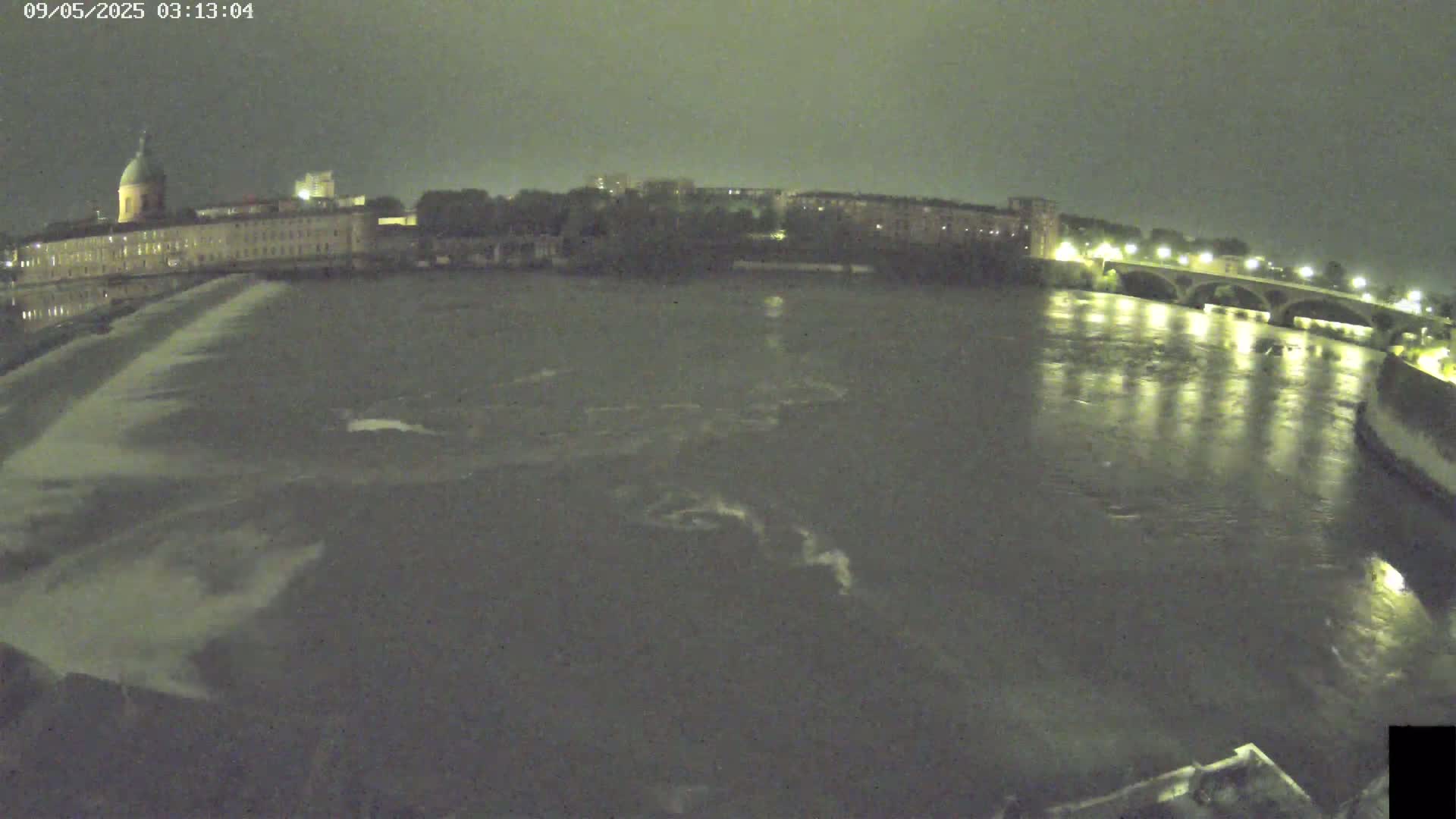 A nighttime view of a river flowing past a large building with a dome, a bridge, and other buildings along the riverbank under a dark, cloudy sky.