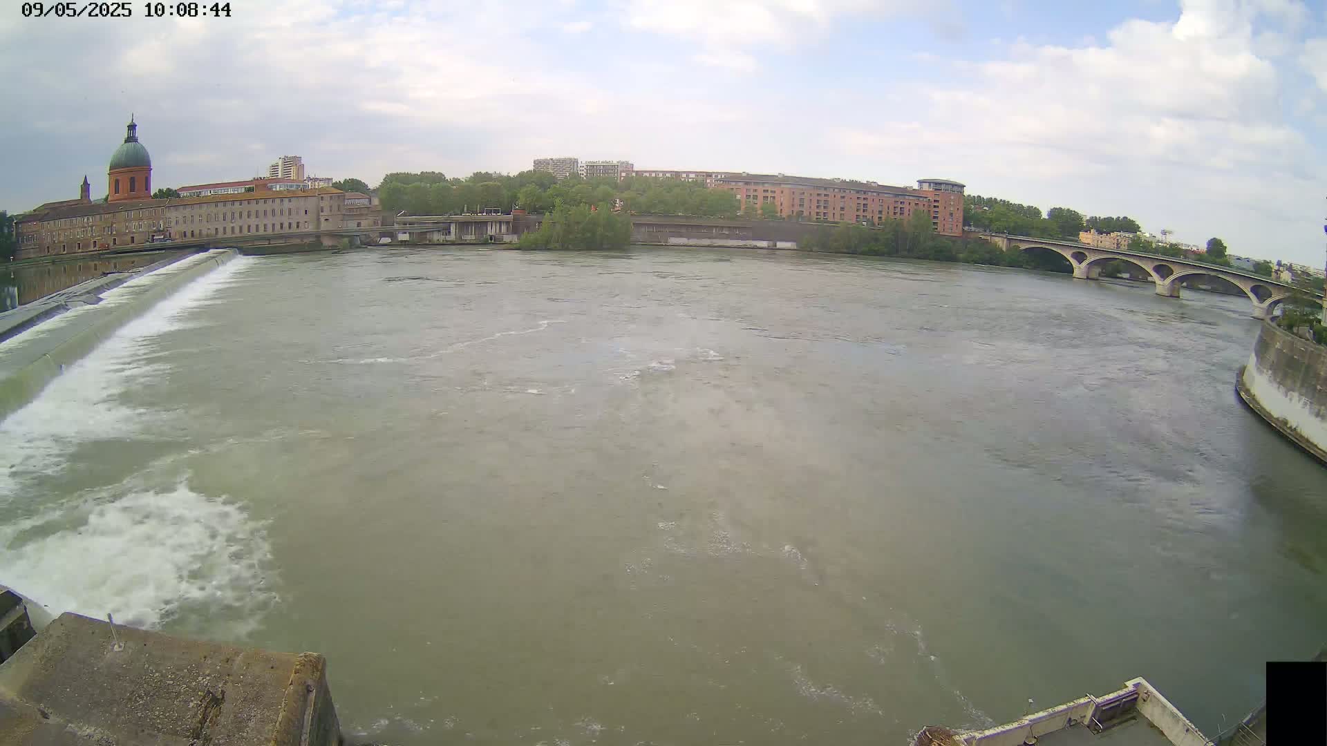 A wide shot of a river flowing past a dam and under a bridge, with buildings and trees visible on the banks under a partly cloudy sky.