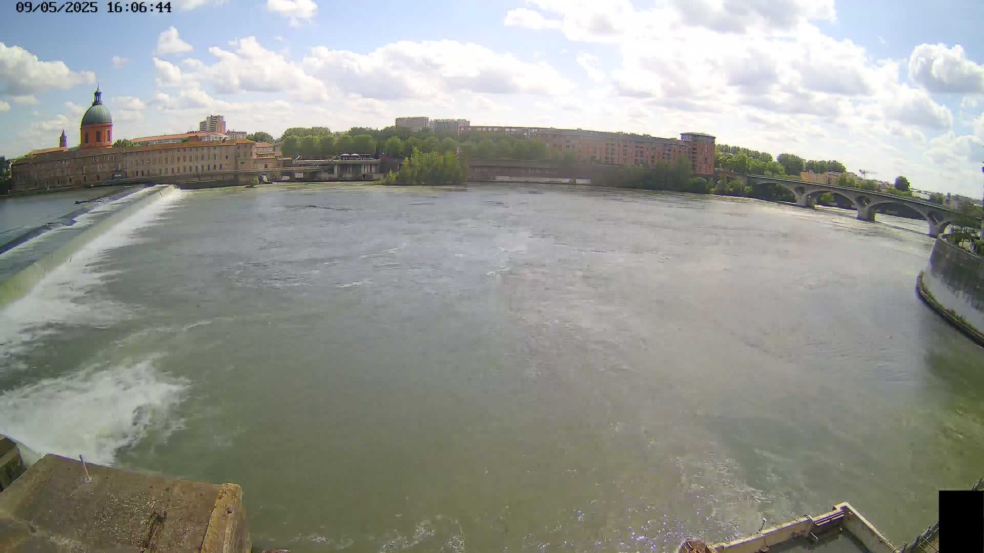 A wide river flows under a partly cloudy sky past a dam, a large building with a dome, and a bridge.