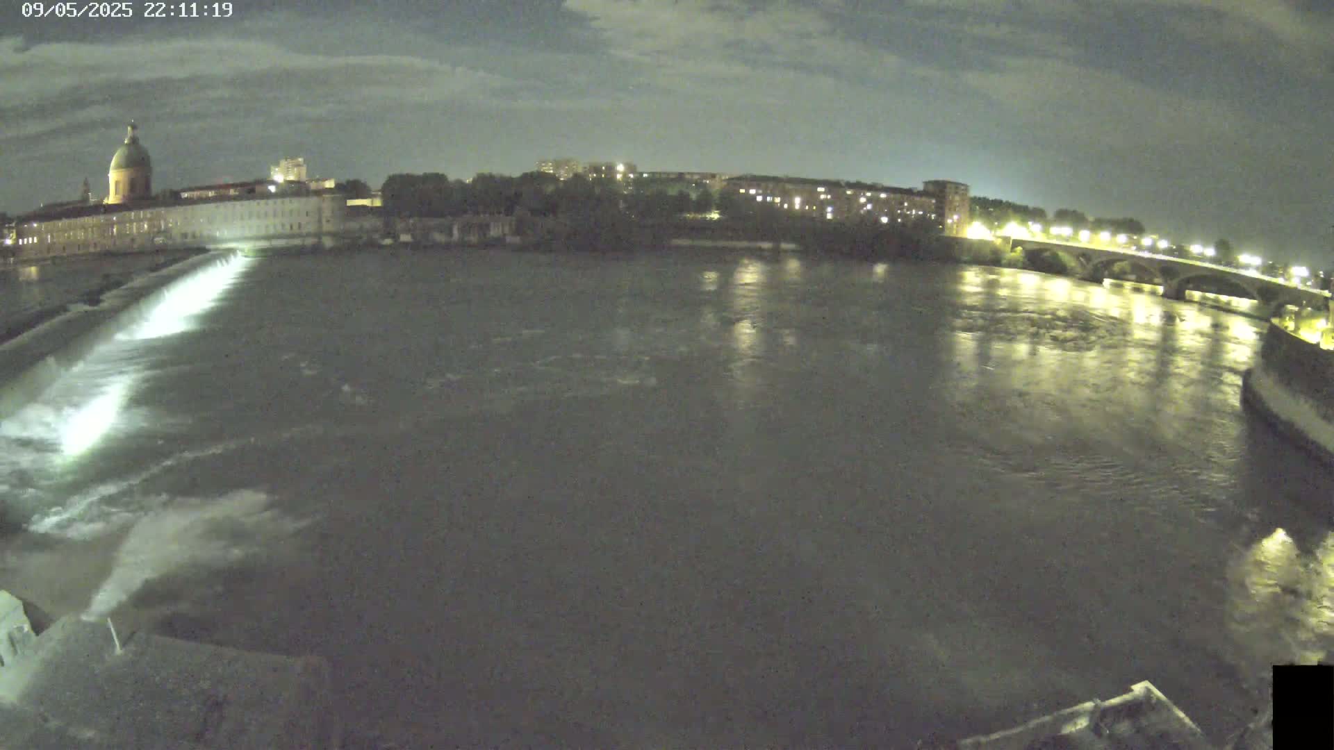 A nighttime view of a river flowing past a city with a domed building, a dam, and a bridge, under a partly cloudy sky.
