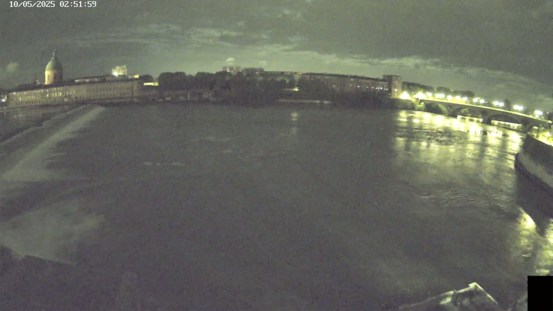 A nighttime view of a river flowing past a city with buildings and a bridge under a cloudy sky.