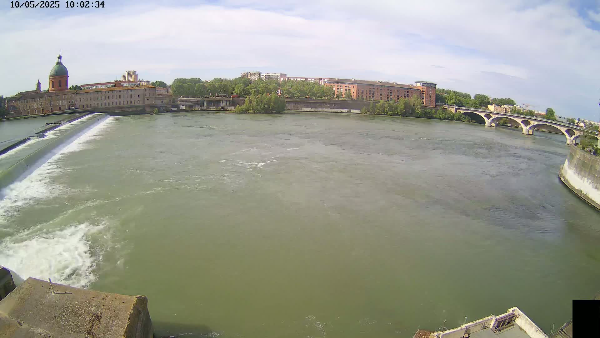 A wide shot shows a river flowing past a dam and under a bridge on a partly cloudy day, with buildings visible along the riverbanks.