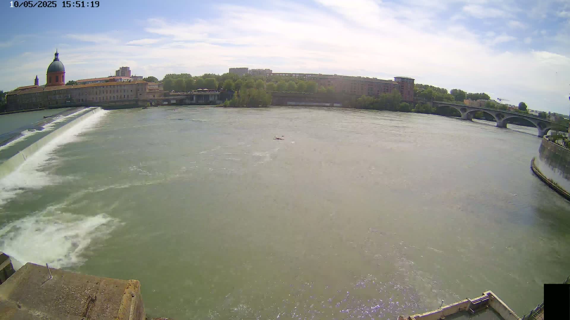 A wide shot shows a river with a dam and a bridge under a mostly sunny sky; a small boat is visible in the center of the river.