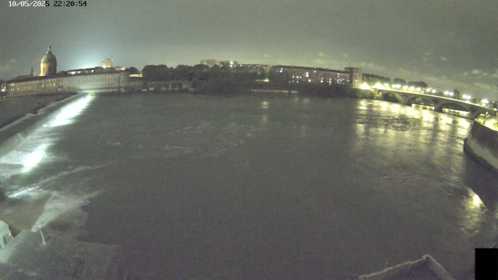 A nighttime view of a river flowing past a city with a large building and bridge, under a dark, overcast sky.