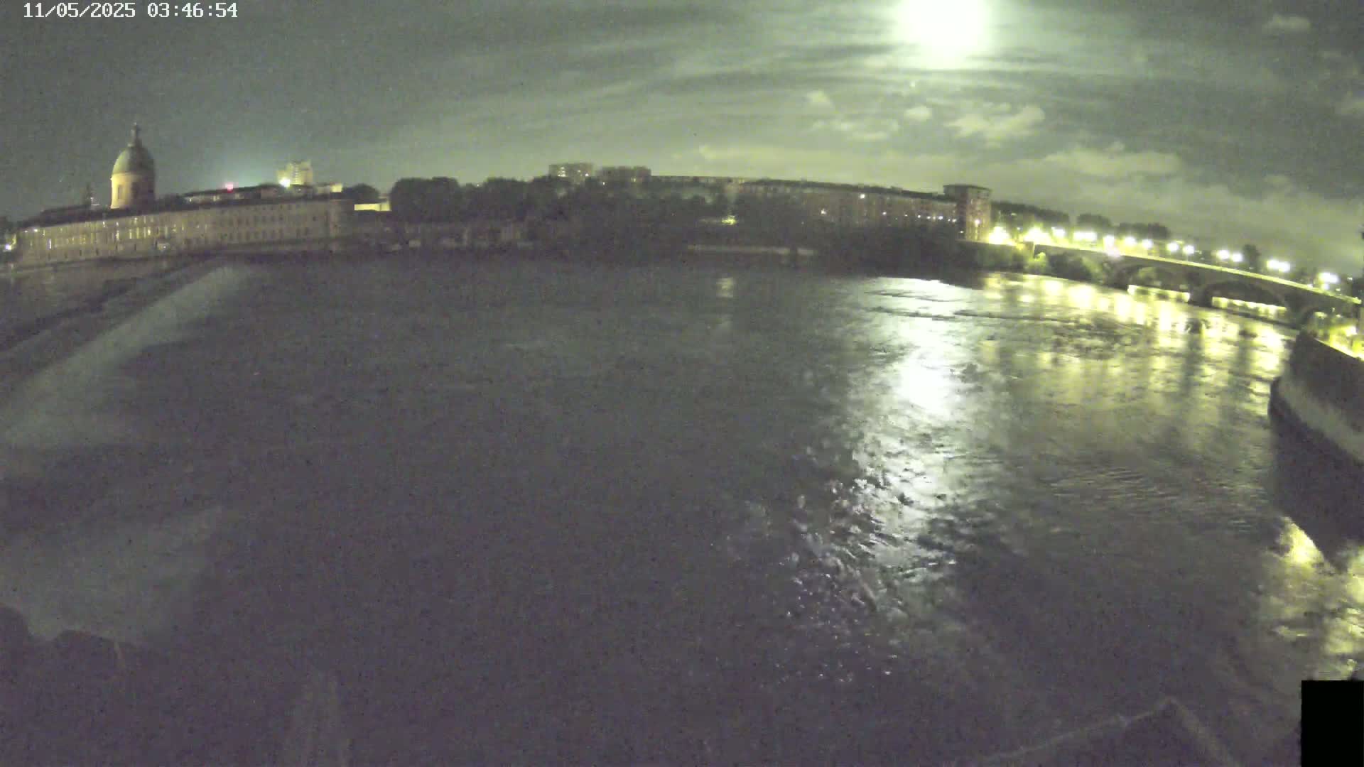 A nighttime view of a wide river reflecting the light from a bridge and buildings on the far bank under a partly cloudy sky.