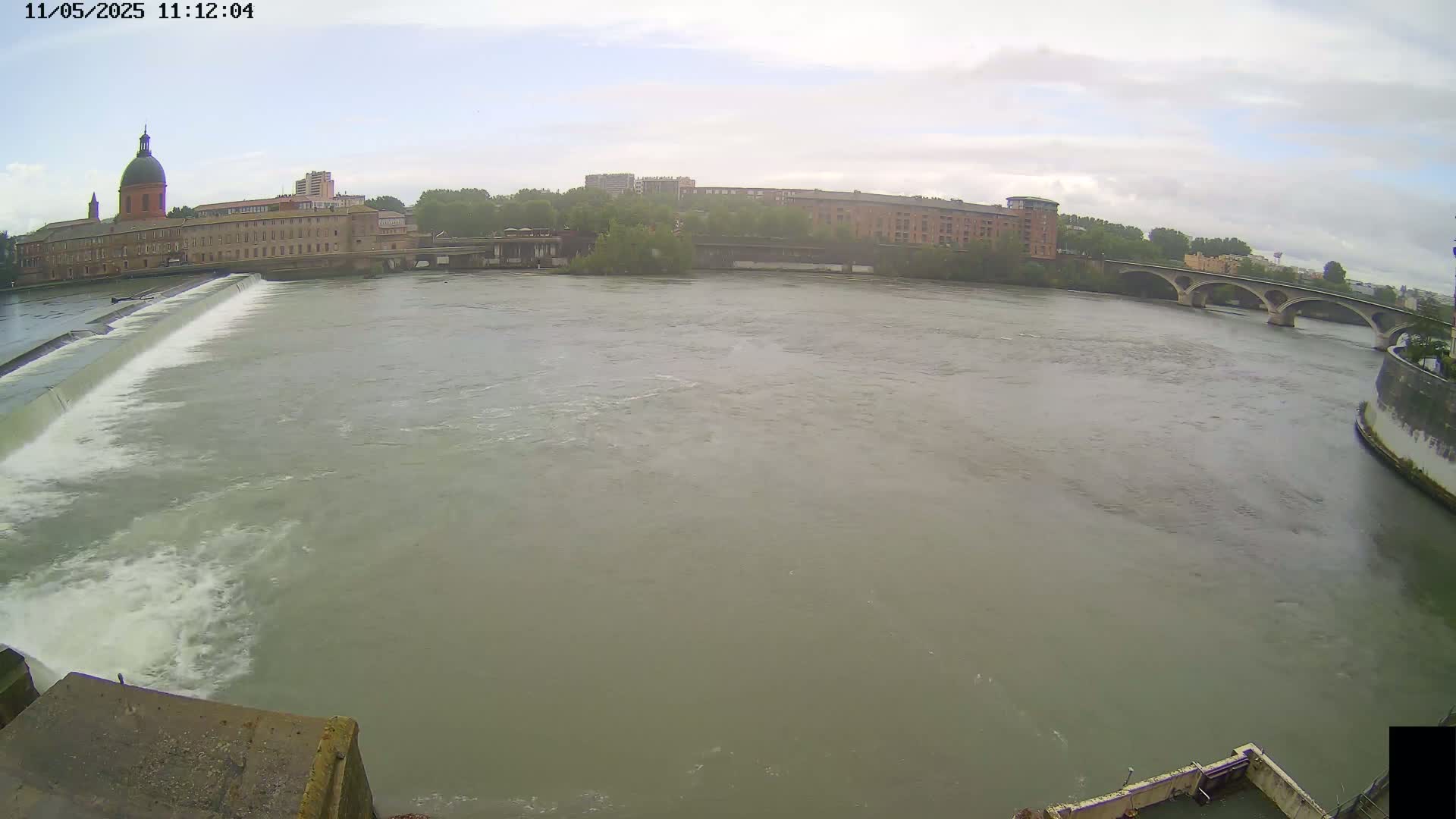 A wide river flows past a dam and under a bridge, with overcast skies and buildings lining the riverbanks.