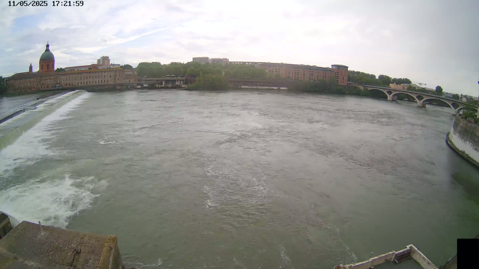 A wide shot shows a river flowing past a dam and under a bridge on an overcast day, with buildings visible on the far bank.
