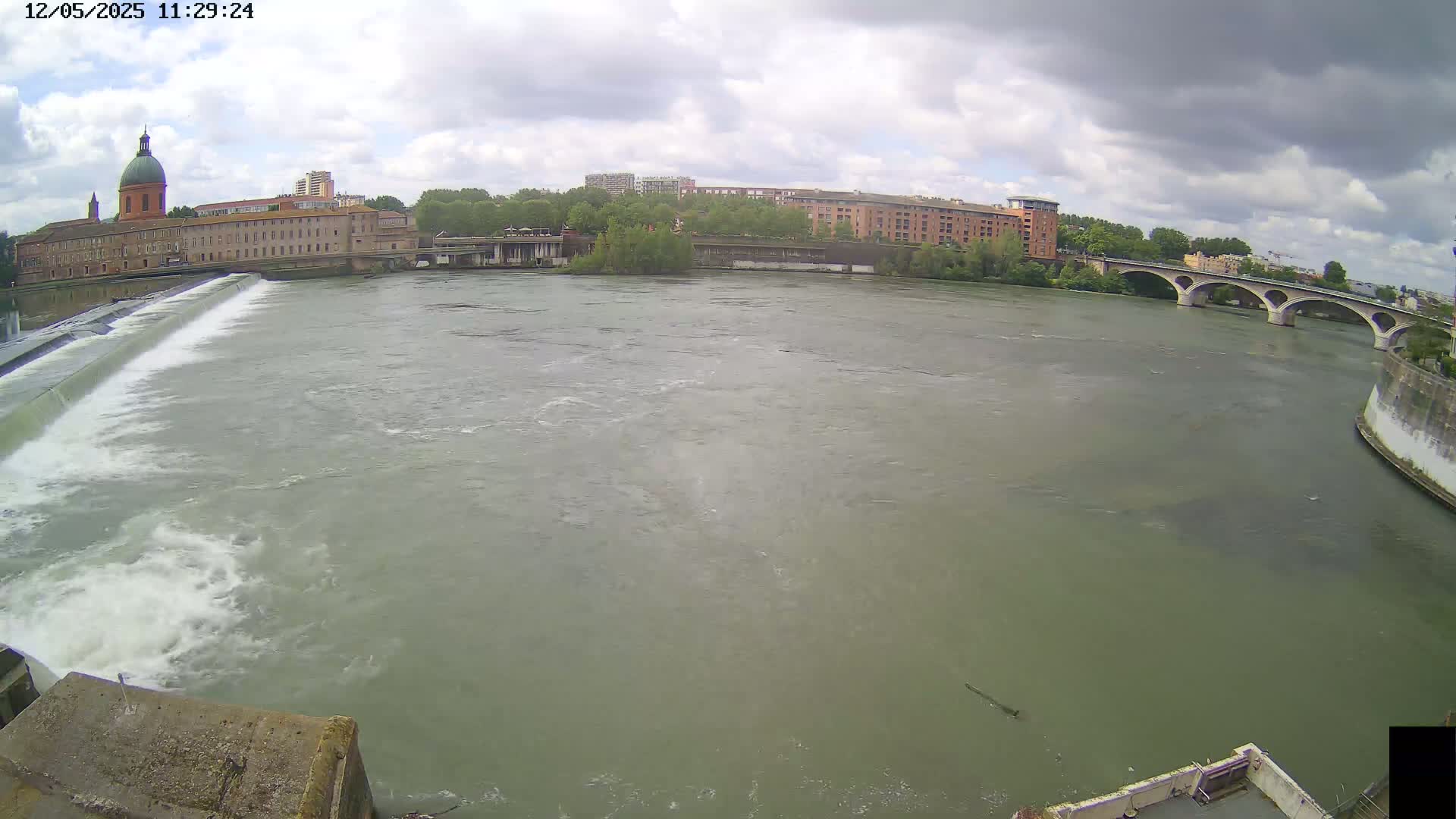 A wide shot of a river flowing past a dam and under a bridge, with overcast skies and buildings visible in the background.