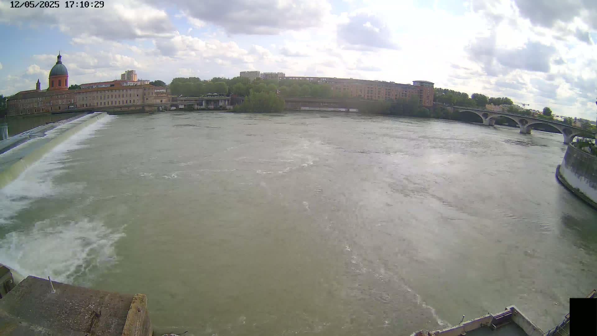 A wide shot shows a river flowing under a bridge, past buildings including one with a dome, on a partly cloudy day.