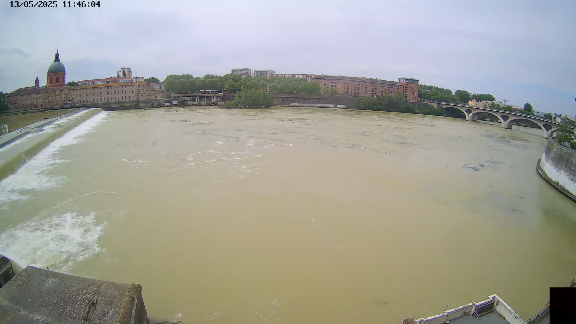 A wide, muddy river flows past a city with buildings, a bridge, and a dam under an overcast sky.