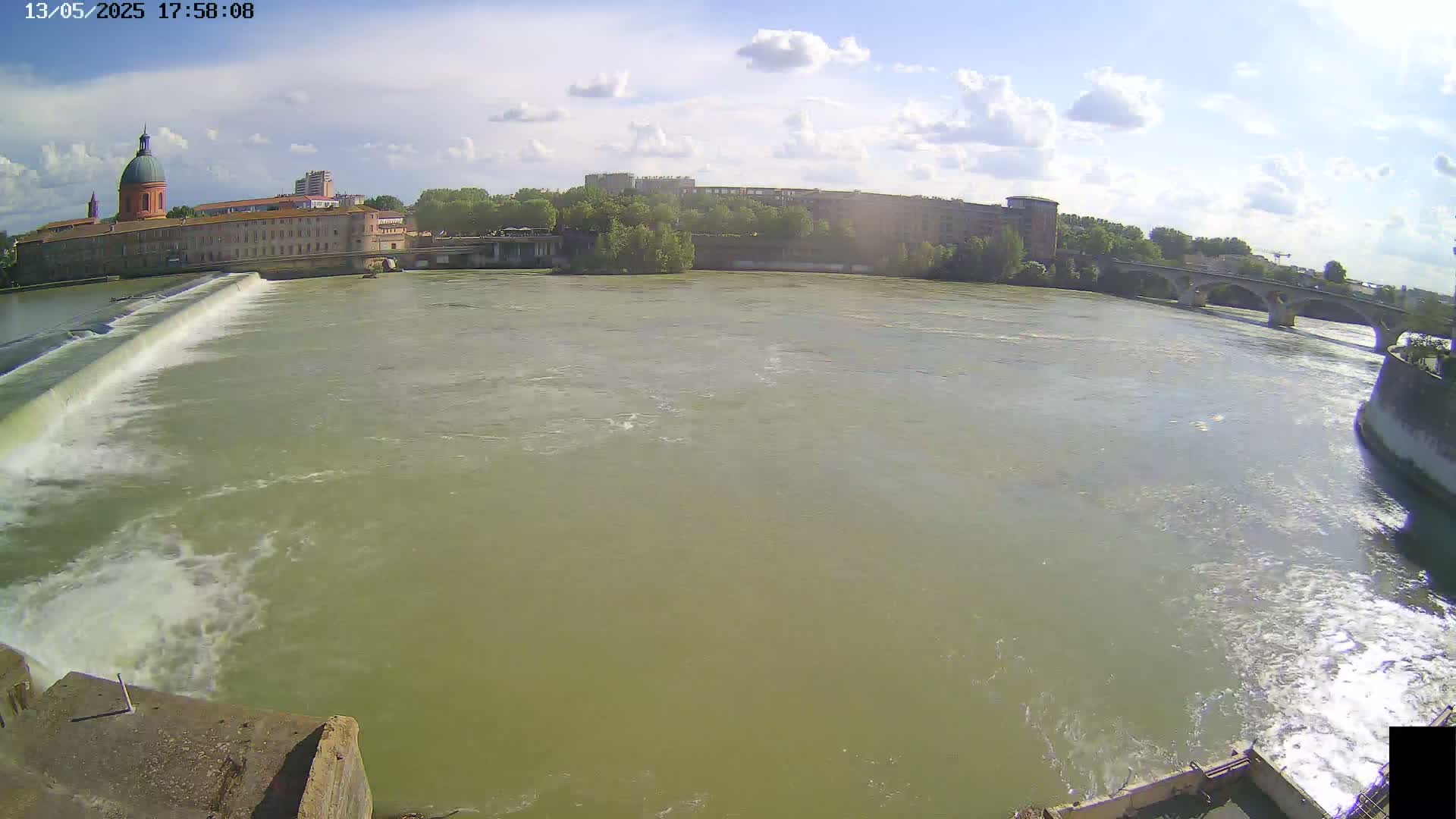 A wide, murky river flows under a partly sunny sky past a large building with a dome, other buildings, and a bridge.