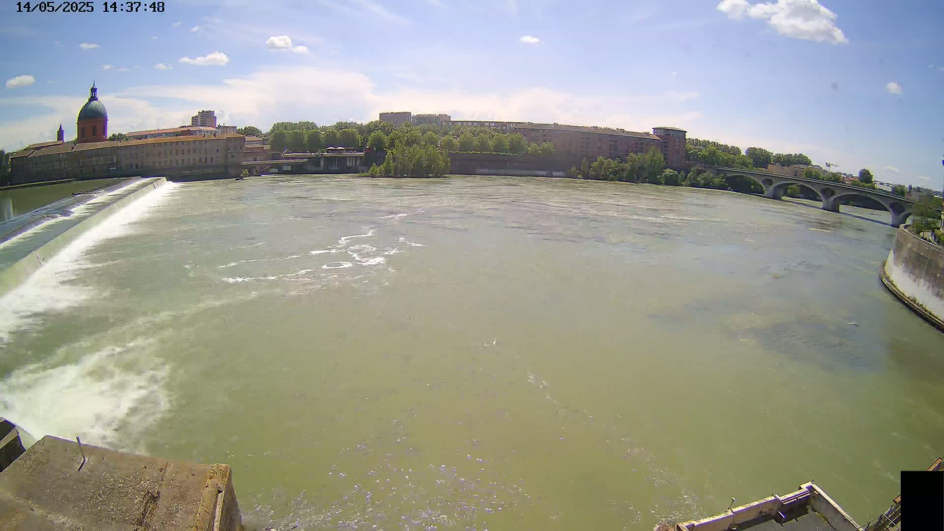 A wide shot shows a river flowing past a dam and under a bridge, with buildings and a domed structure visible in the background on a sunny day with some clouds.