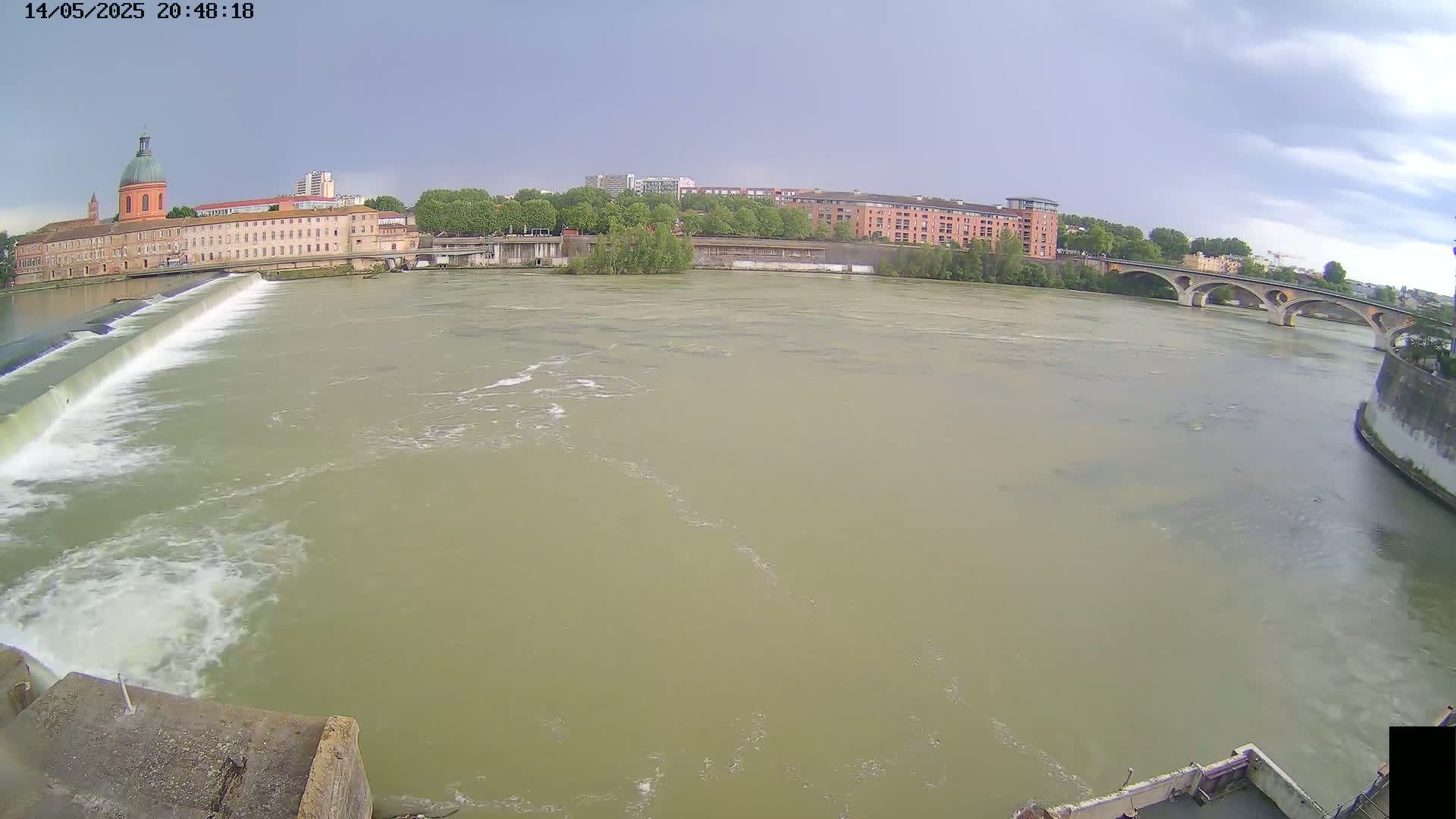 A wide, muddy river flows past a dam and under a bridge, with buildings and trees visible on the far bank under a partly cloudy sky.