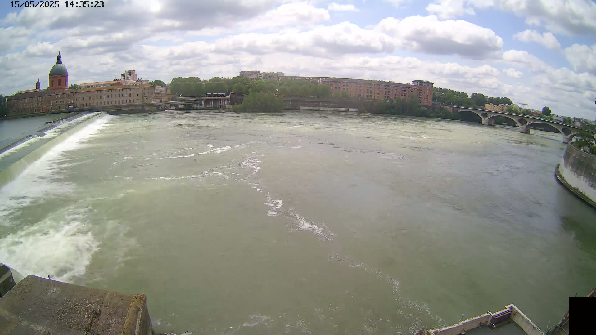 A wide shot of a river flowing under a bridge, past a dam and buildings, on a partly cloudy day.