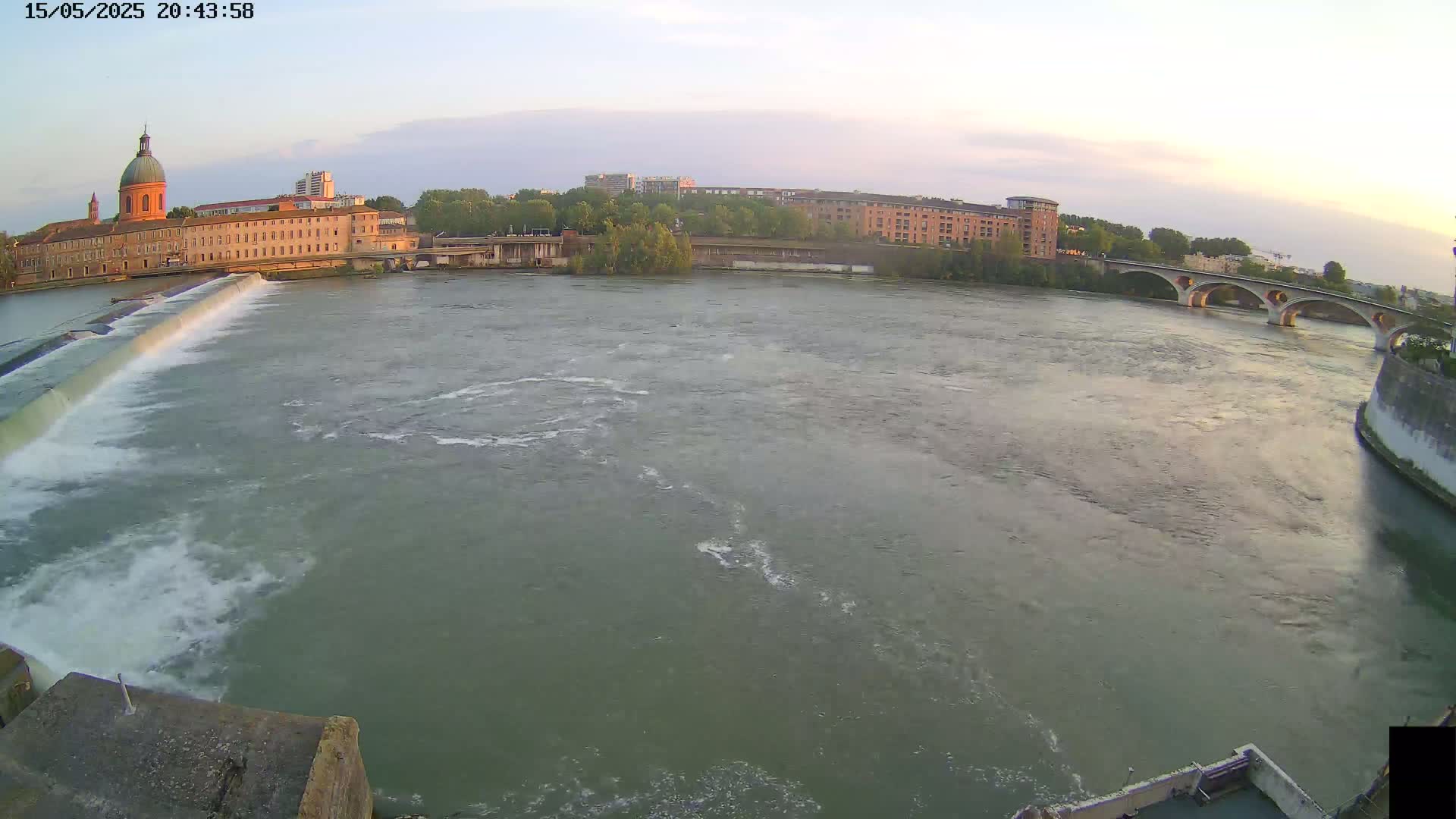 A wide, calm river flows past a dam and under a stone bridge, with buildings and trees visible on the shore under a twilight sky.
