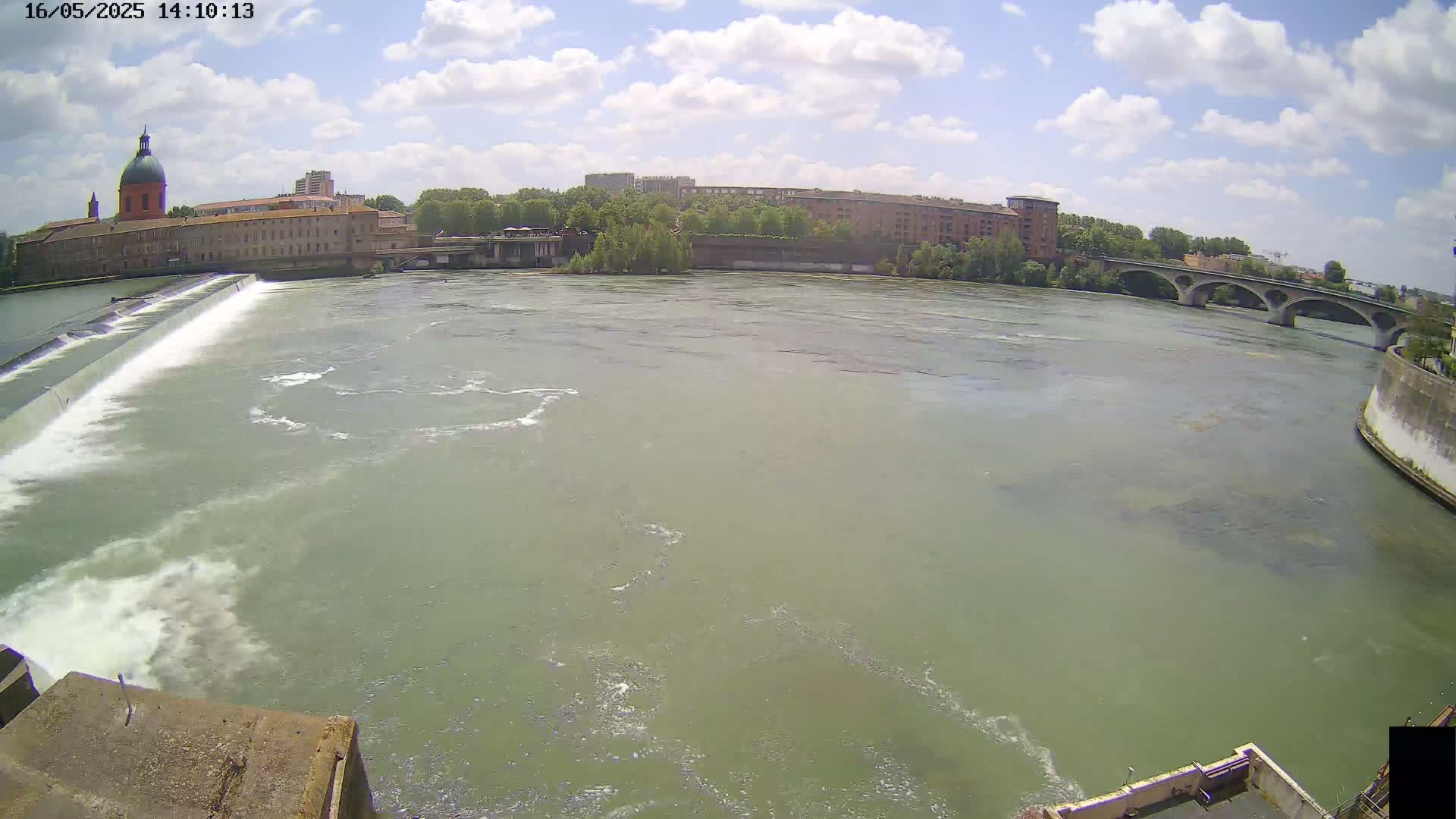 A wide shot of a river flowing under a bridge on a partly cloudy day, with a dam and buildings visible in the background.
