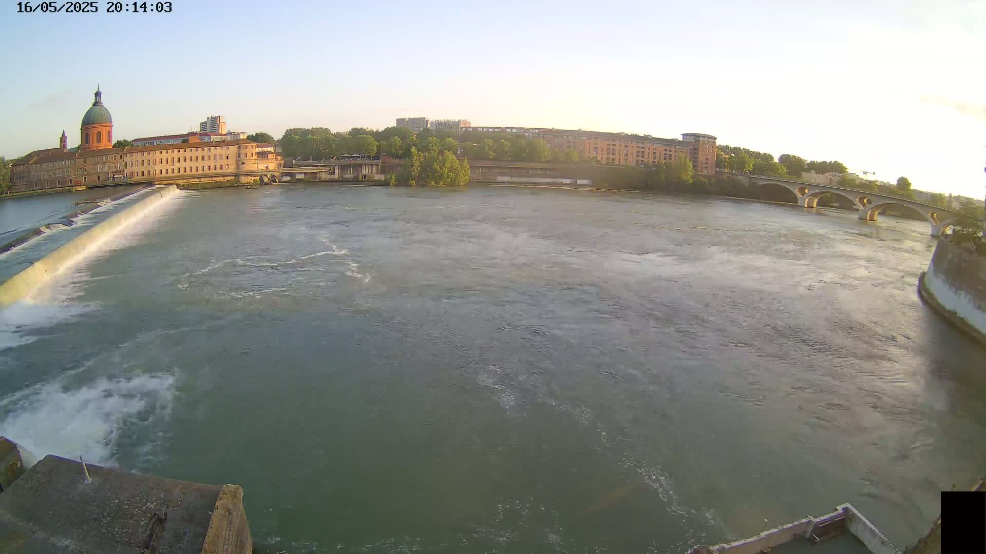A river flows past a city with buildings, a dam, and an arched bridge under a clear evening sky.