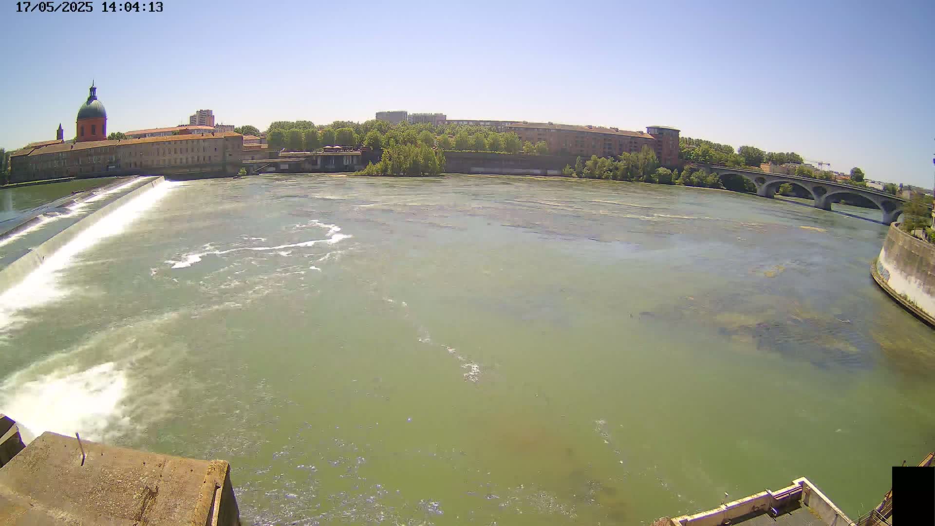 A wide shot on a sunny day shows a river flowing past a dam, with buildings and a bridge visible in the background.
