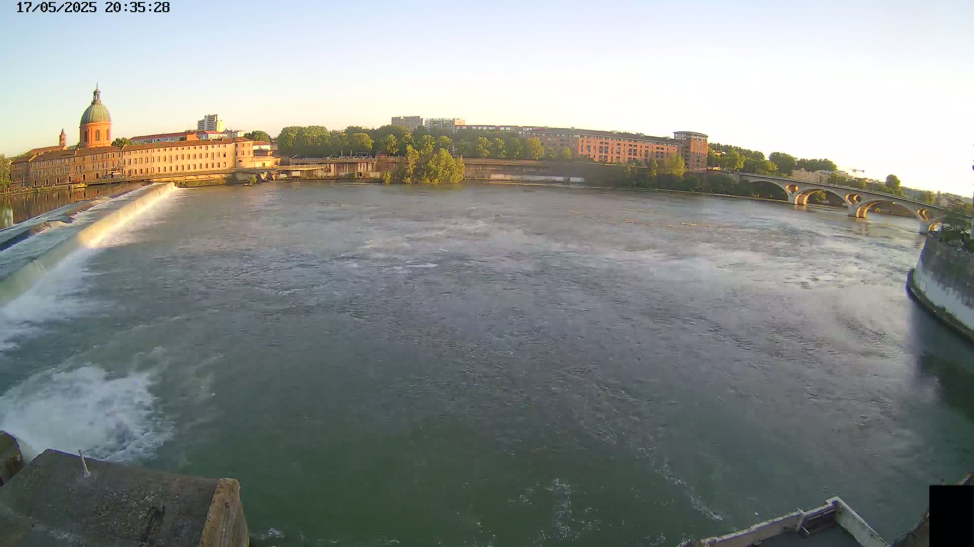 A wide shot of a river with a dam and a bridge under a clear, evening sky, featuring buildings along the riverbank.