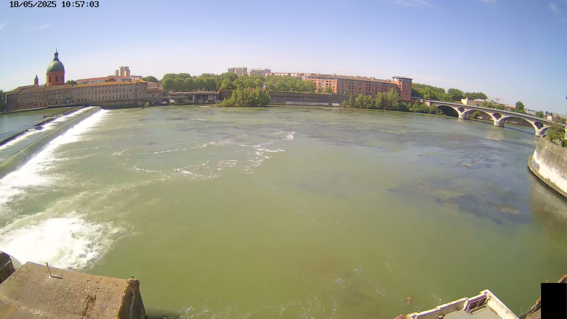 A wide shot of a river flowing under a bridge on a sunny day, with a dam and a large building visible in the background.