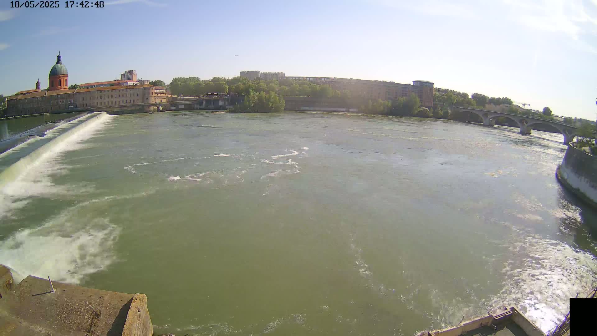 A wide shot of a river flowing past a dam and buildings under a clear sky.