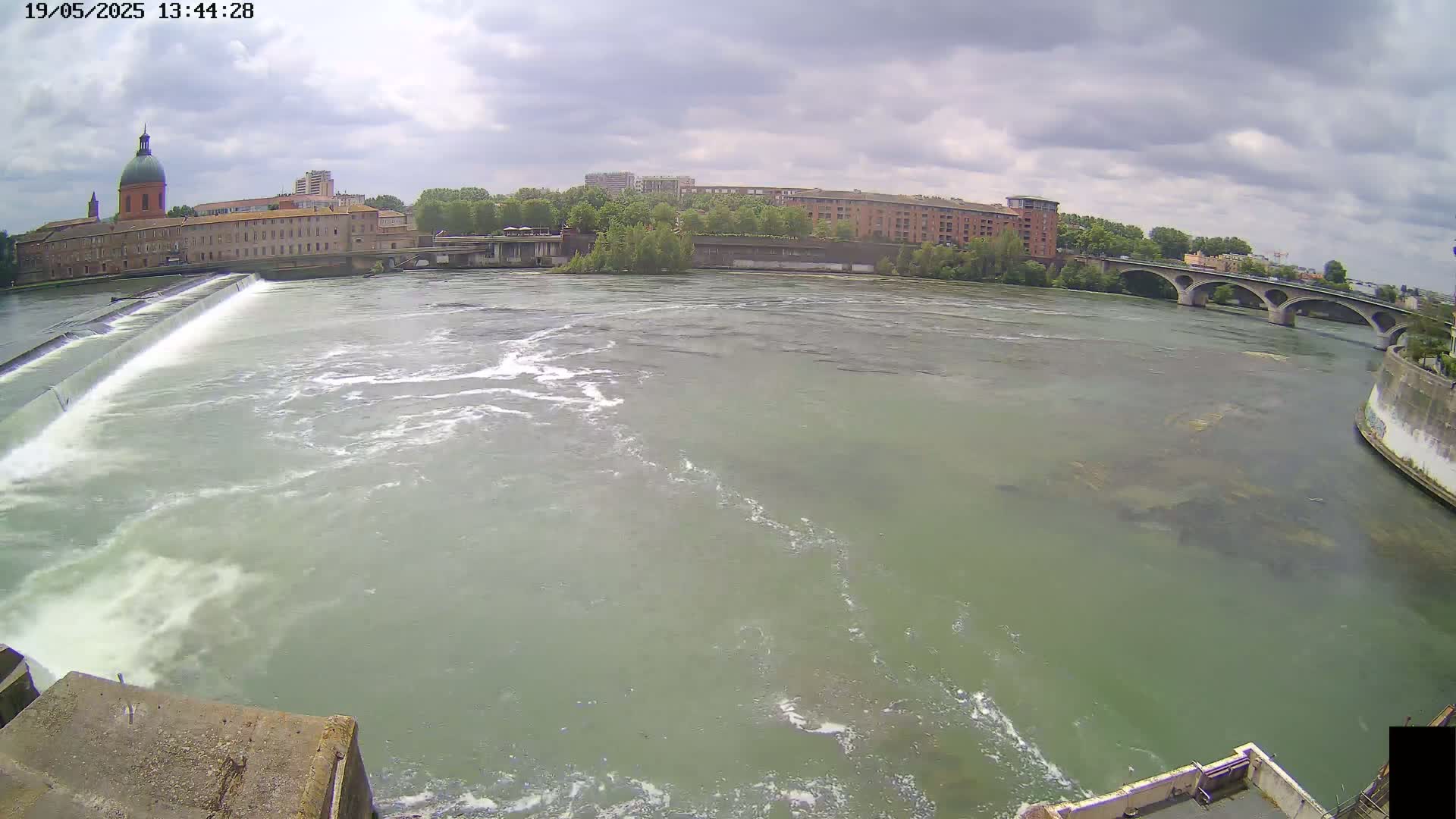 A wide shot of a river flowing past a dam and under a bridge, with buildings and cloudy skies in the background.