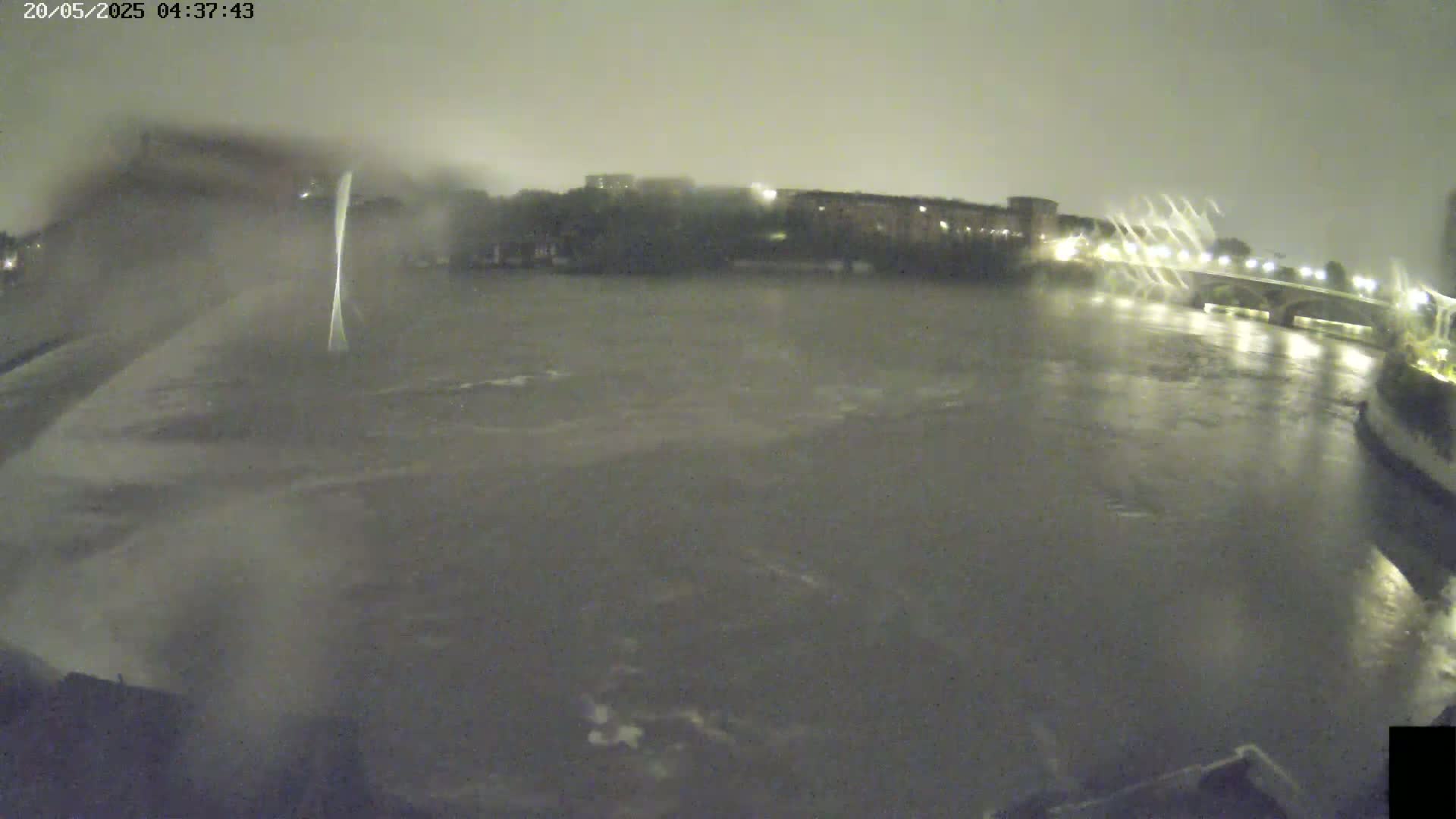 A nighttime view of a rain-swept river flowing under a bridge, with buildings dimly visible on the far bank.
