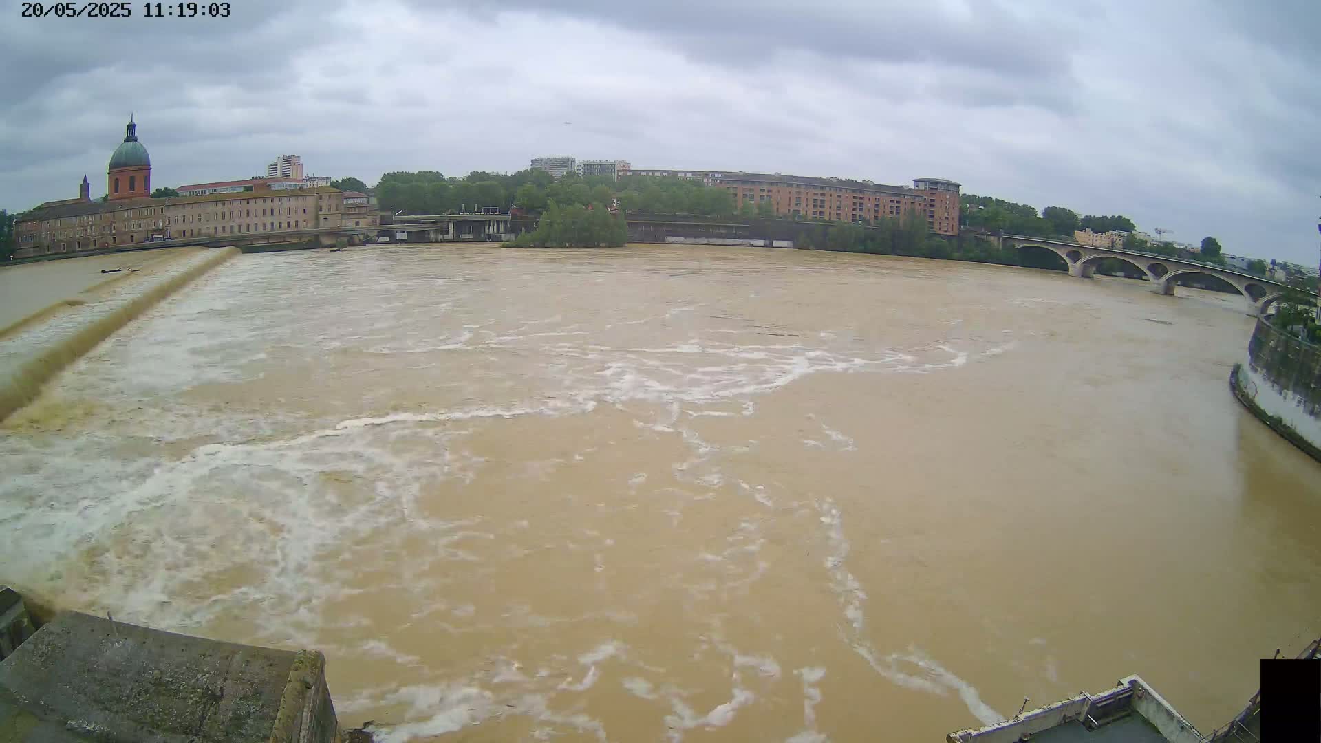 A muddy, rapidly flowing river surges past buildings and a bridge under an overcast sky.