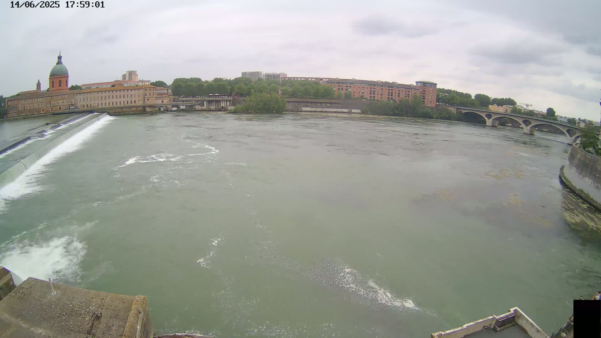A wide river flows past a dam and buildings under an overcast sky.