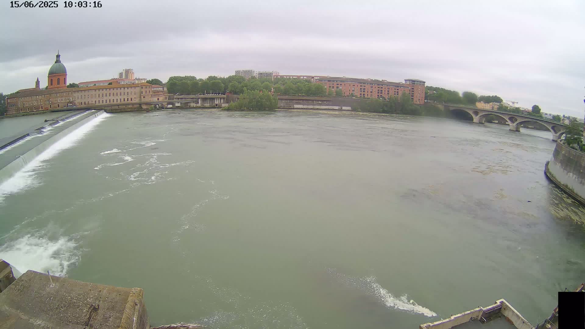 A wide shot of a river flowing past a dam and under a bridge on an overcast day, with buildings and trees visible along the riverbanks.