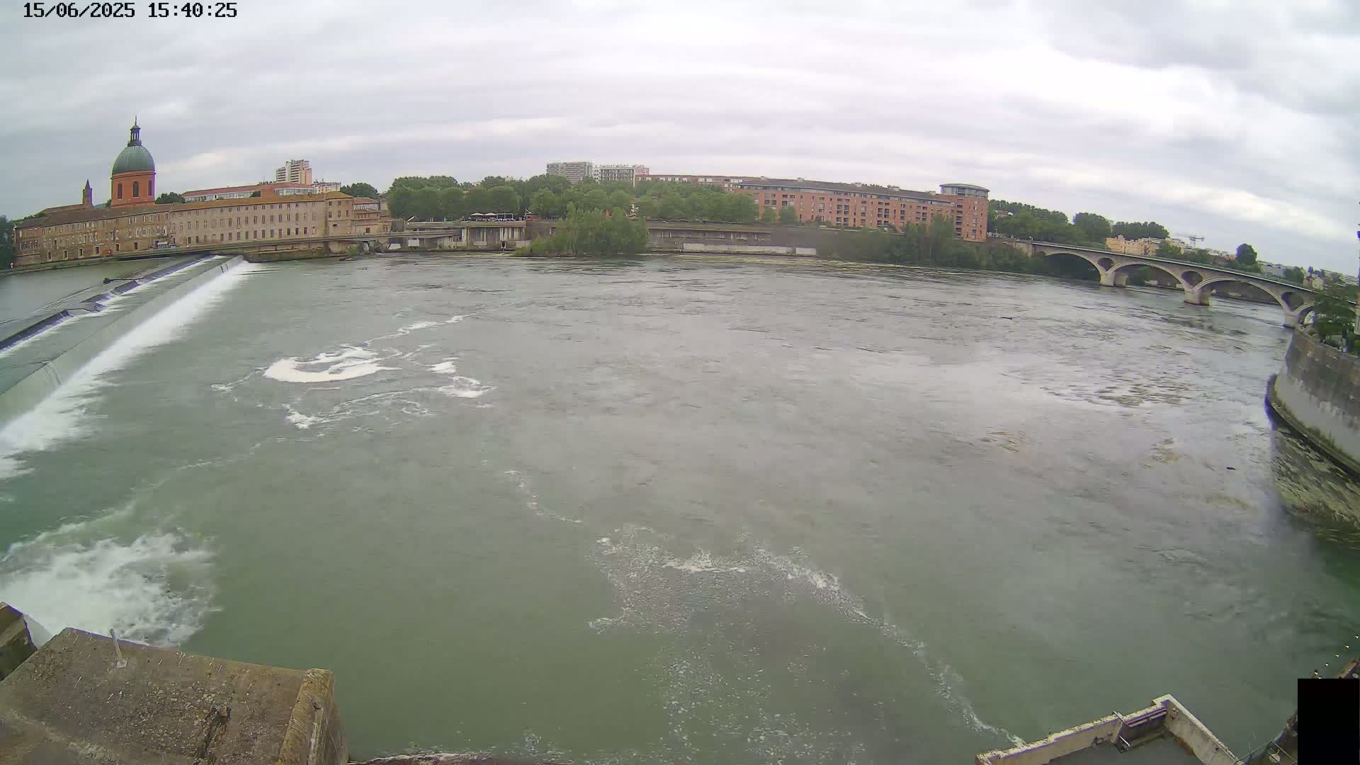 A wide shot shows a river flowing under a bridge on an overcast day, with a dam and buildings visible in the background.