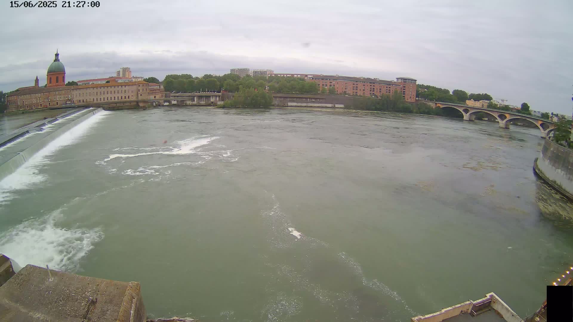 A wide shot shows a river flowing under a bridge on an overcast day, with a dam and buildings visible in the background.