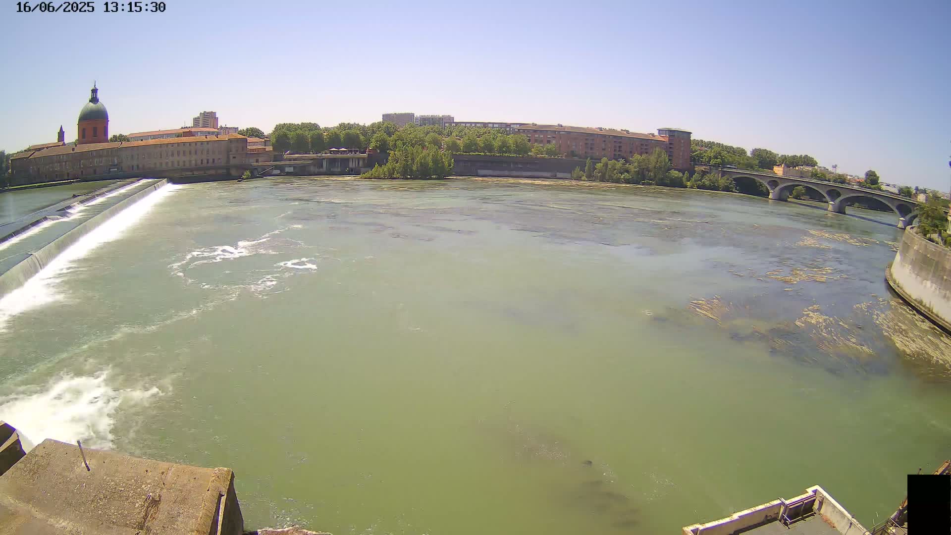 A wide shot shows a river flowing past a dam and under a bridge on a sunny day, with buildings and trees visible on the banks.