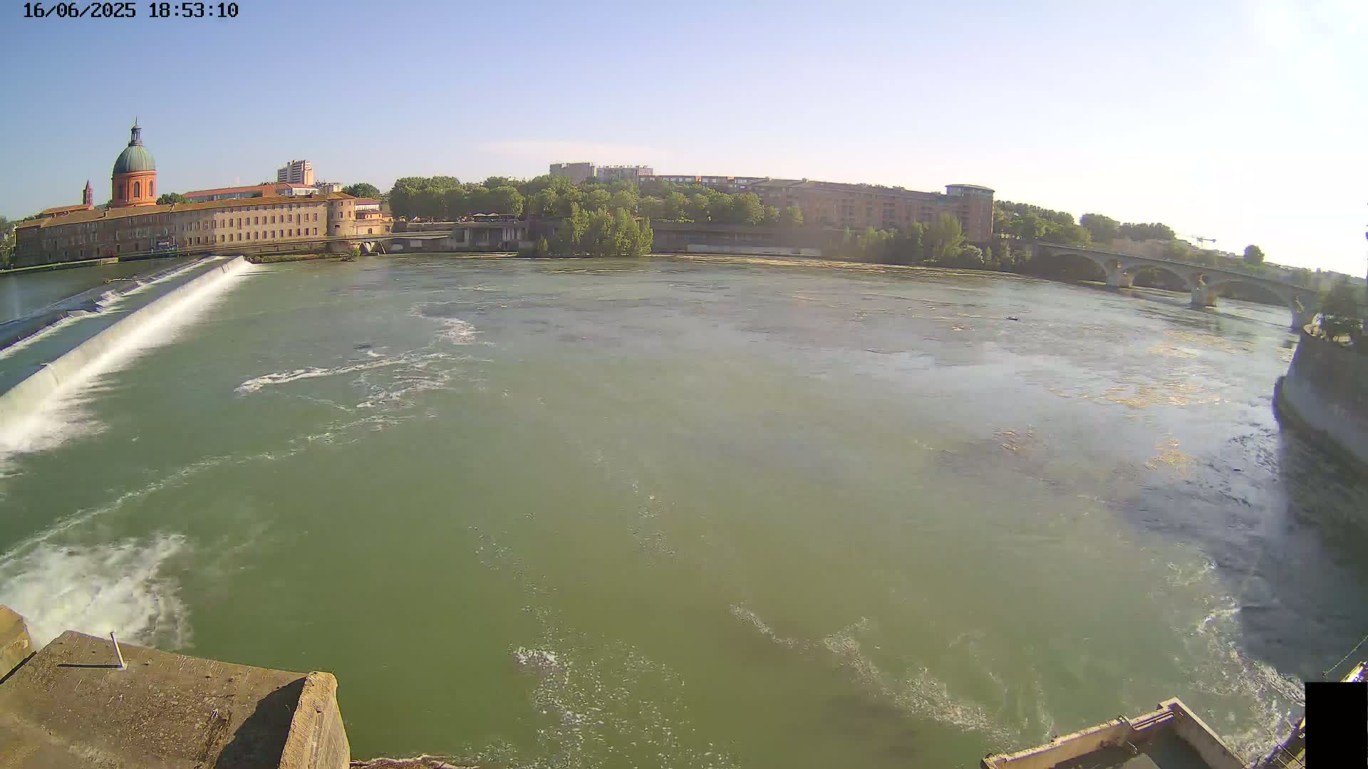 A wide shot of a river flowing past a dam and buildings under a clear, sunny sky.