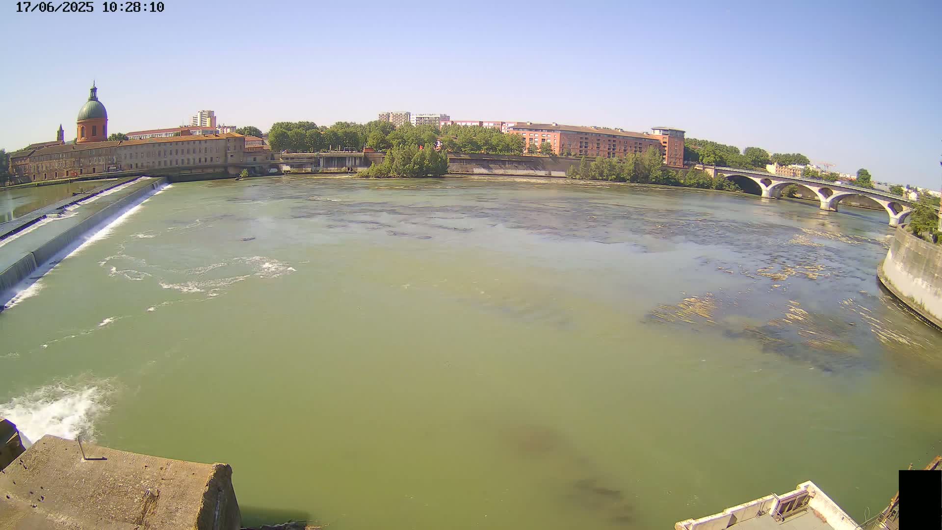 A wide shot on a sunny day shows a river flowing past a dam and under a bridge, with buildings and trees visible on the banks.