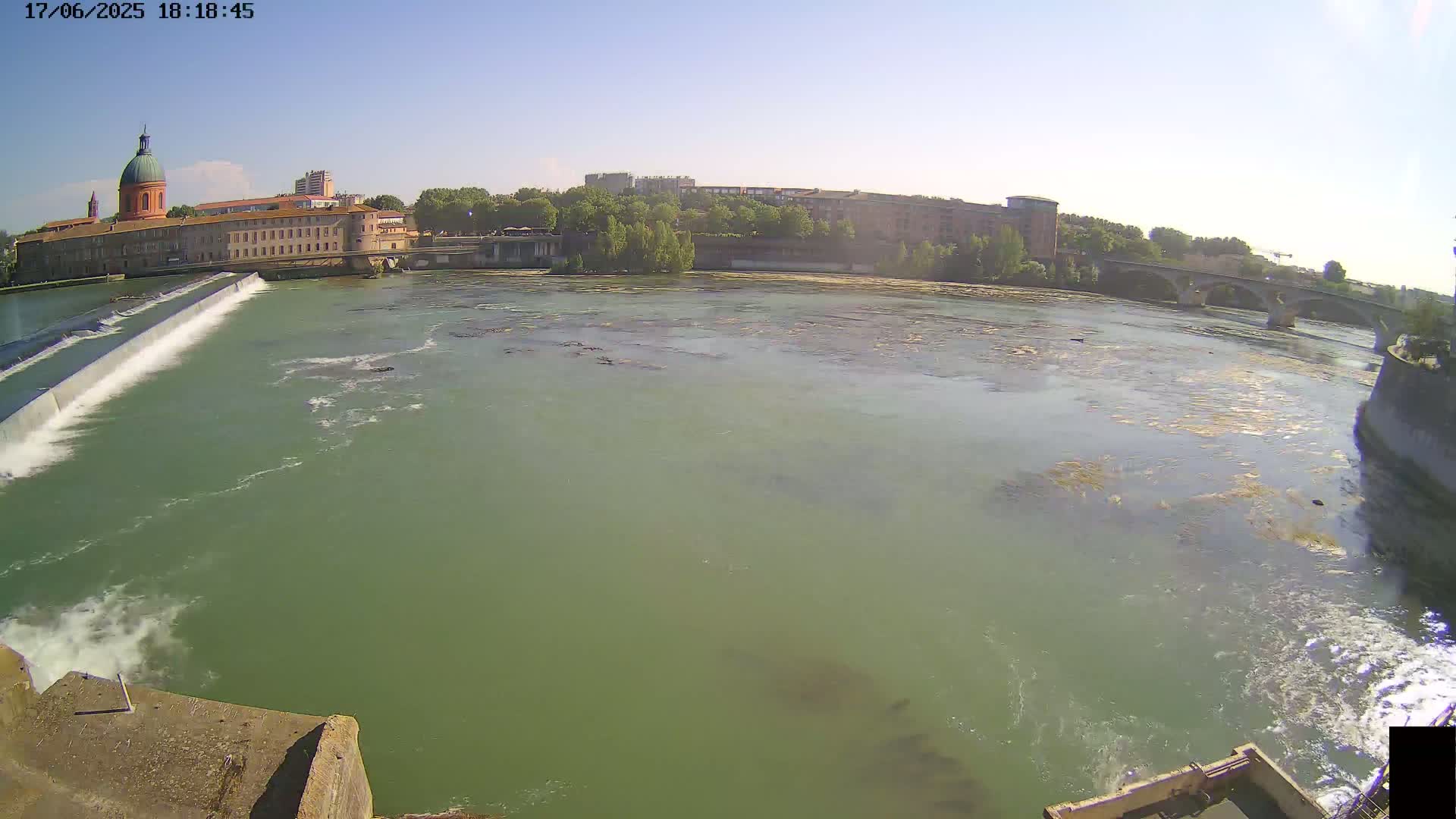 A wide shot shows a river flowing past a dam and buildings on a sunny day.
