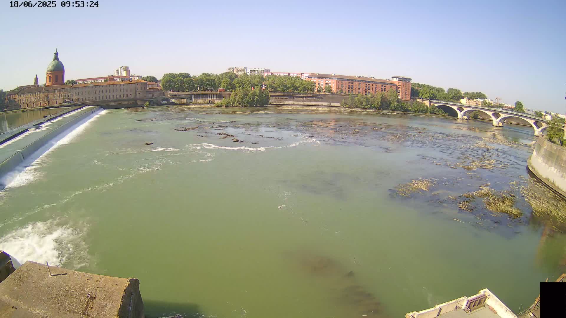 A wide shot shows a river flowing past a dam and under a bridge on a sunny day, with buildings and trees lining the riverbanks.