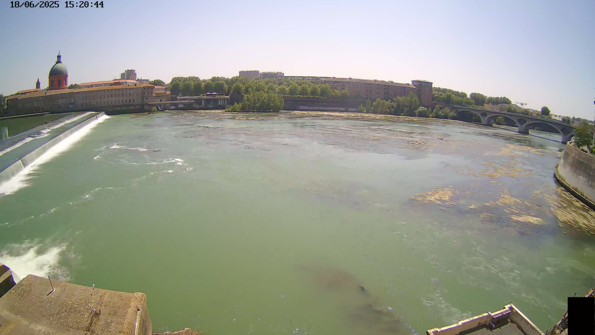 A wide shot of a river flowing past a dam and buildings under a clear sky.
