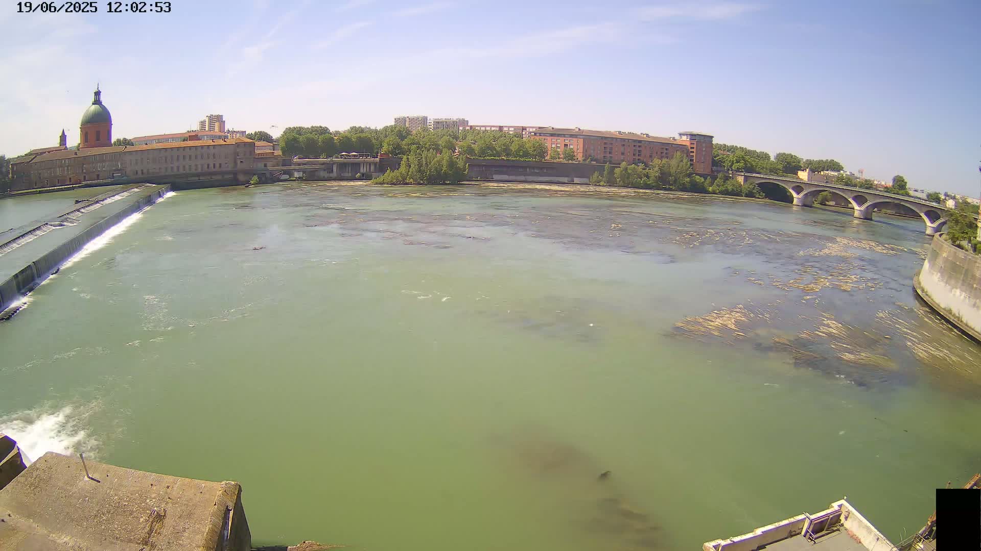 A wide, greenish-brown river flows past a stone dam and under a bridge on a sunny day, with buildings and trees lining the banks.