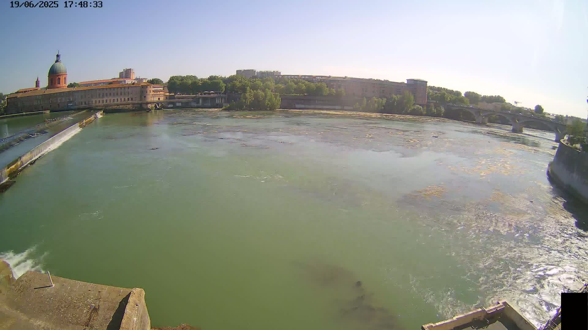 A wide shot of a river flowing past a city under a clear sky, with a dam visible in the foreground.
