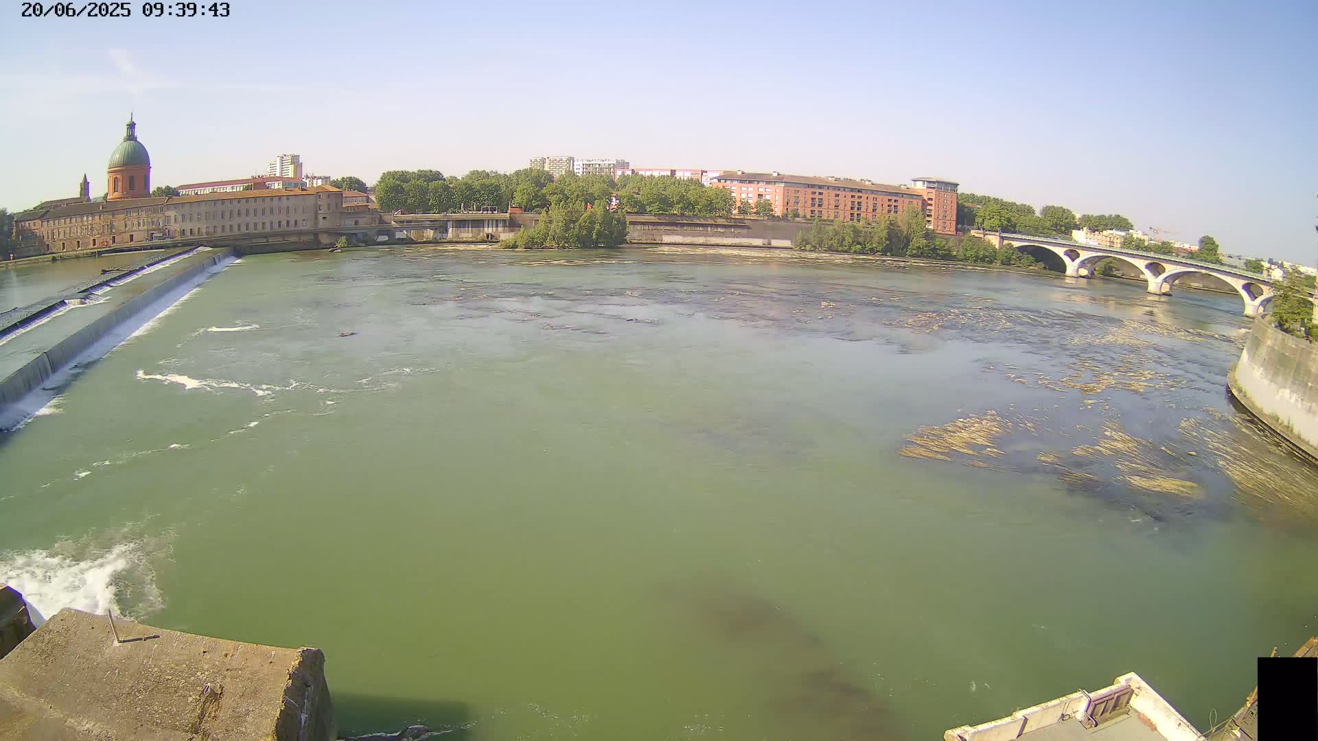 A wide shot of a river with a dam and bridge under a clear, sunny sky, showing buildings and trees along the riverbank.