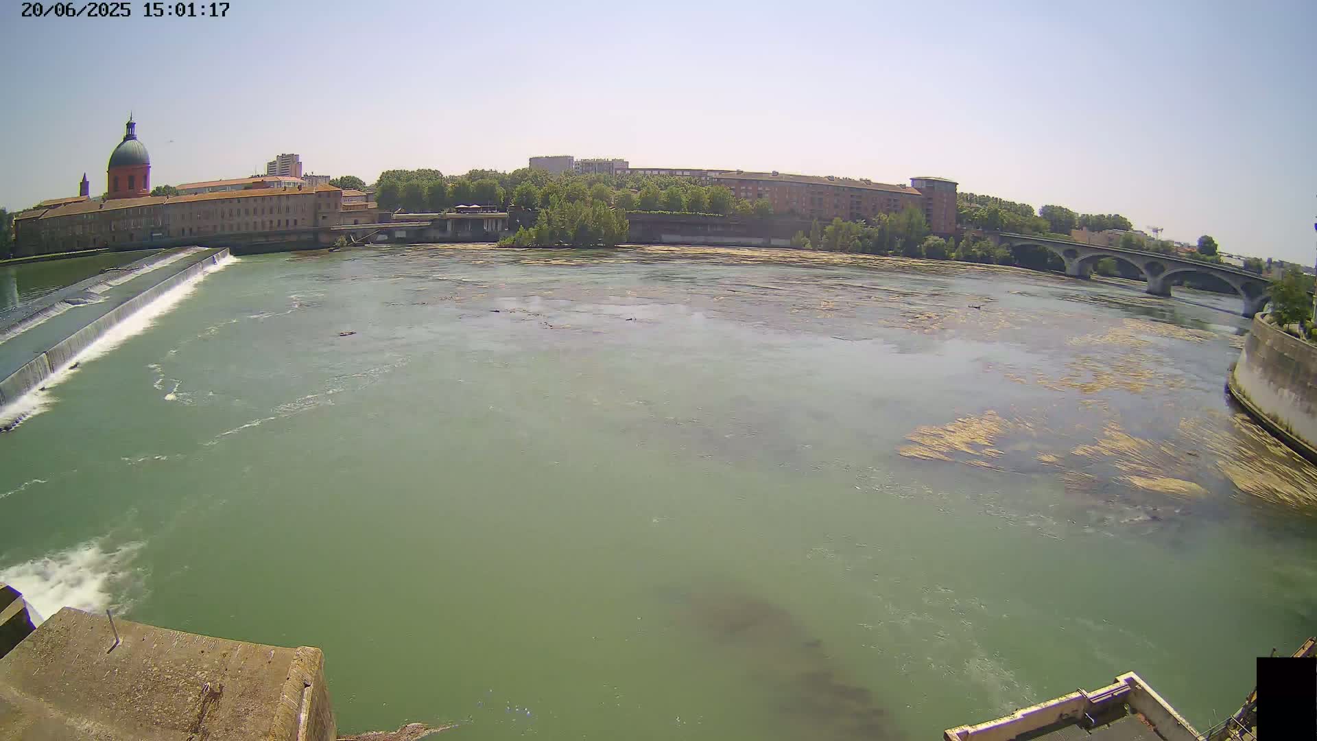 A wide shot of a river flowing past a dam and city buildings under a clear sky.