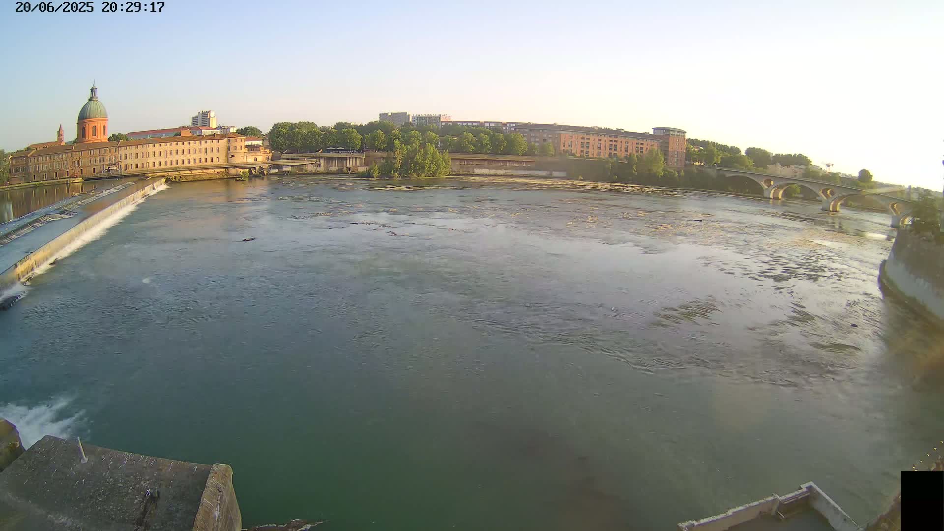 A wide shot of a river flowing past buildings and a bridge under a clear sky.