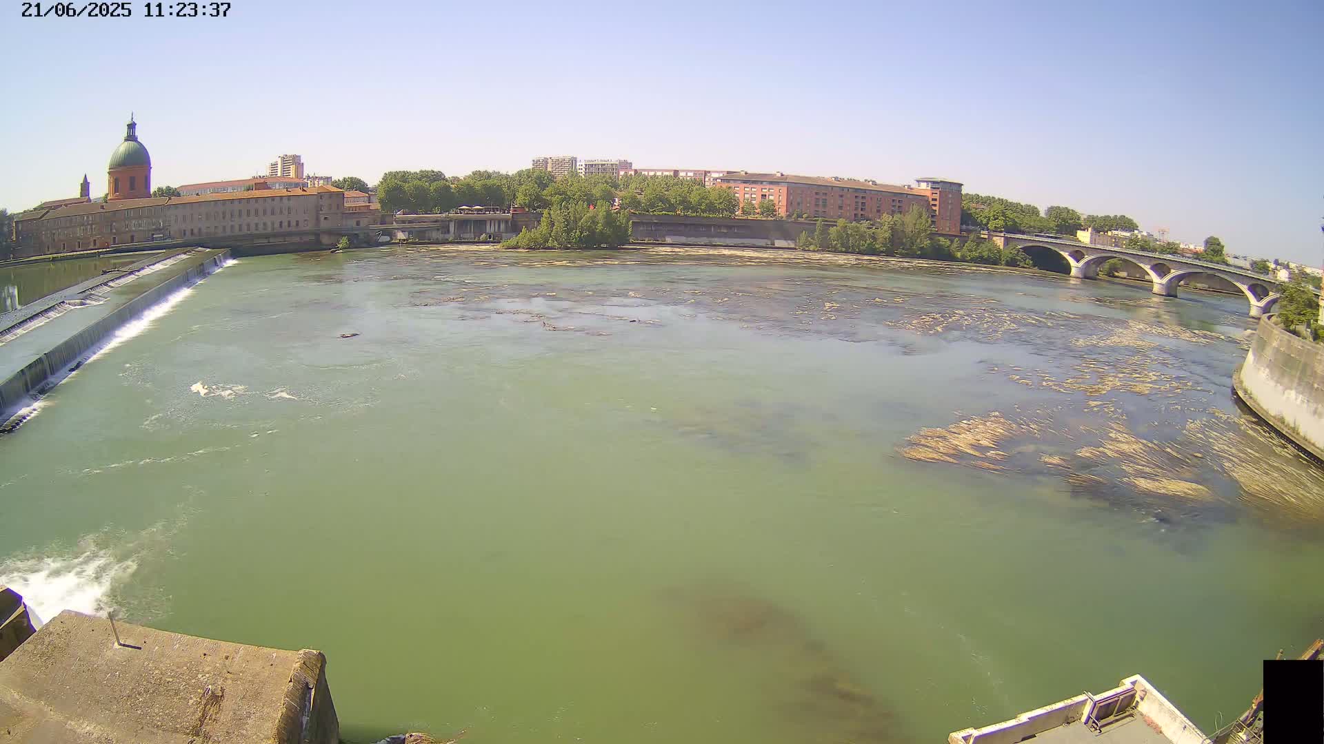A wide shot on a sunny day shows a river flowing past a dam and under a bridge, with buildings and trees visible on either bank.