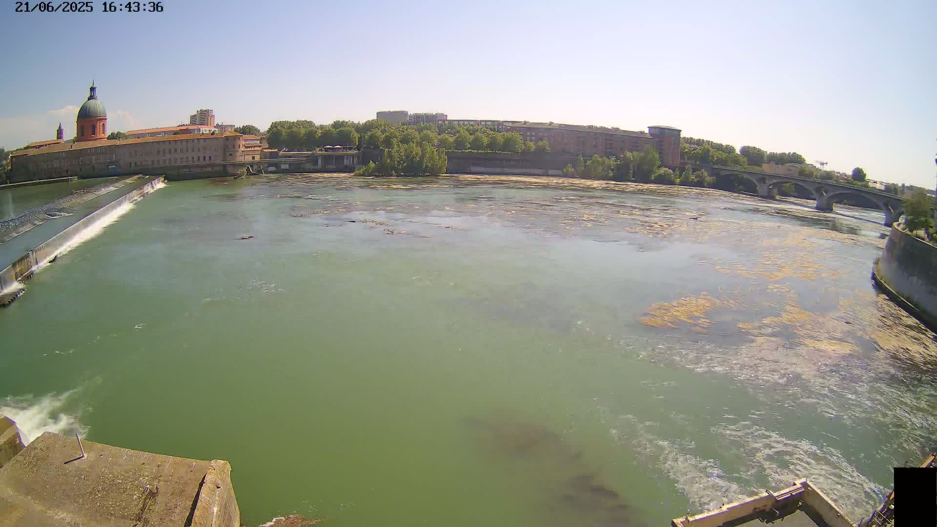 A wide shot of a river flowing past a city on a sunny day, with a dam and bridge visible in the distance.