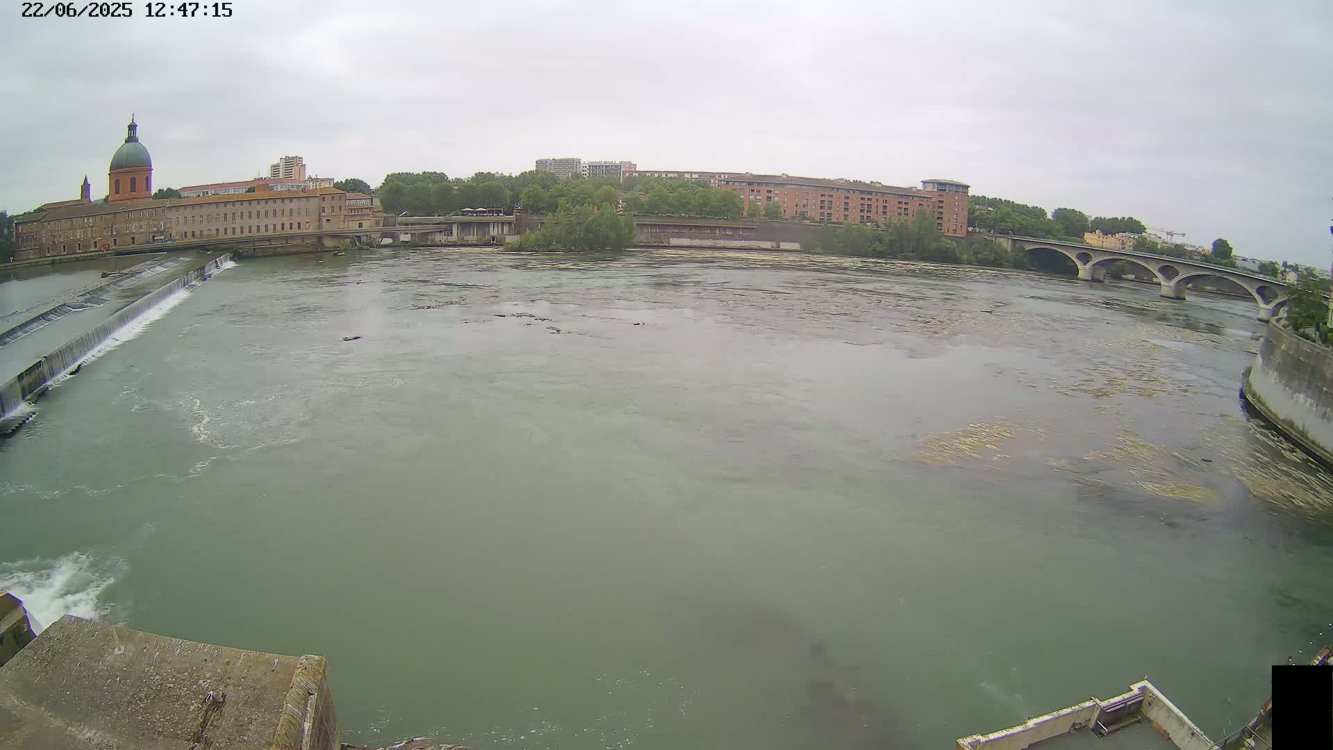 A wide, murky river flows under a bridge and past a building with a dome, on an overcast day.
