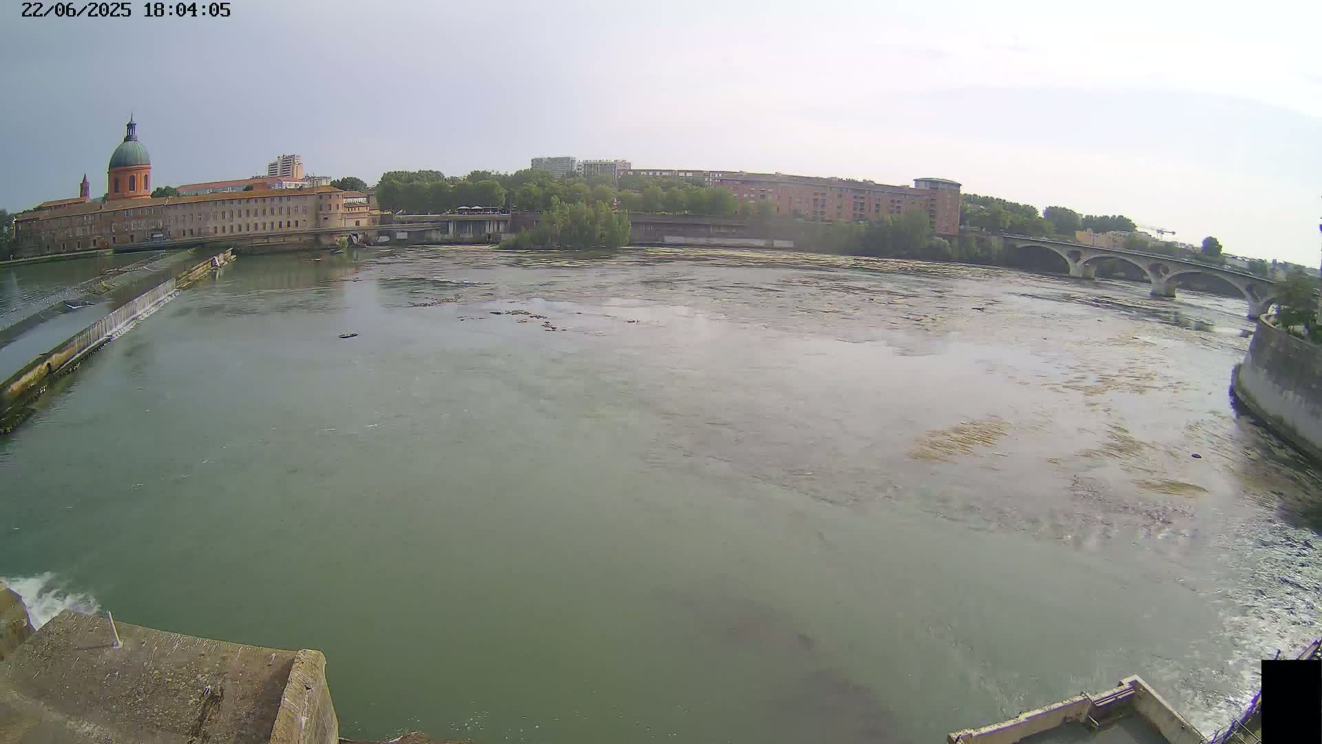 A wide, murky river flows past a large orange-brick building with a dome, a bridge, and other buildings under a mostly clear sky.